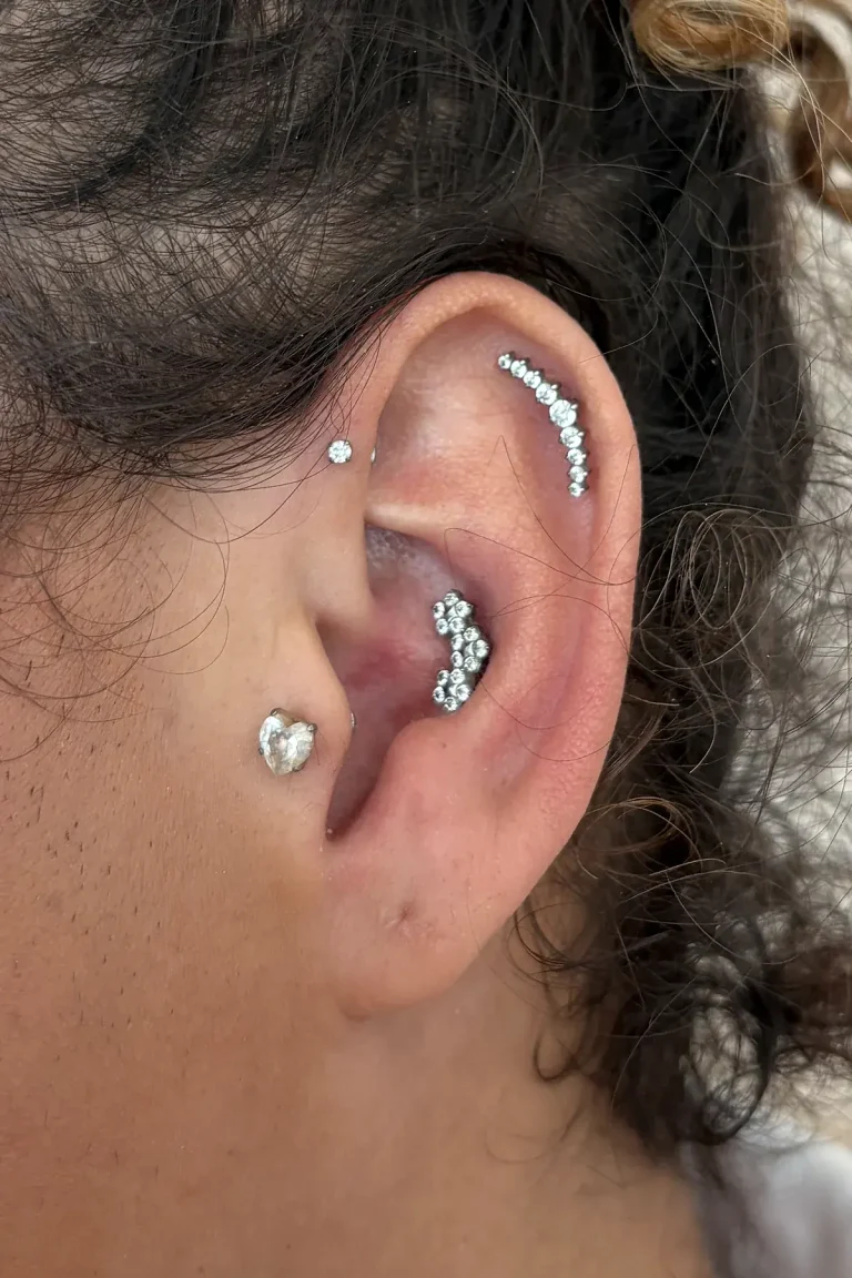 Composição de piercings na orelha: barra curva de zircônias no helix, joia de pedras no conch e pontos de luz na frente da orelha, feita na Visão Tattoo em Curitiba