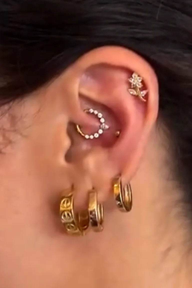 Composição de piercings dourados: daith com argola de zircônias em meia-lua, flor na cartilagem superior e argolas no lóbulo, feita na Visão Tattoo em Curitiba