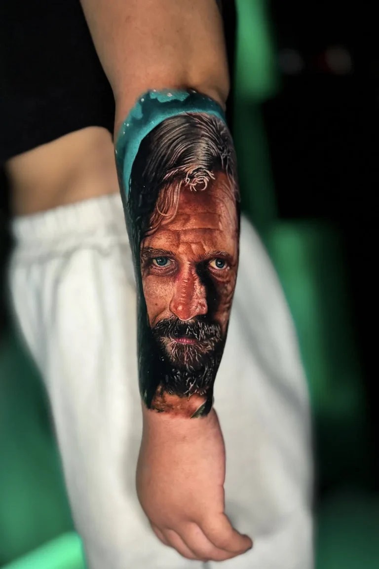 Tatuagem realista colorida no antebraço: rosto de homem de cabelo comprido, barba e olhos azuis, com fundo turquesa, feita na Visão Tattoo em Curitiba