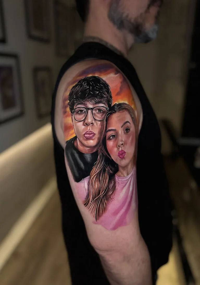 Tatuagem realista colorida no braço: retrato de casal fazendo biquinho com pôr do sol ao fundo, feita na Visão Tattoo em Curitiba